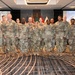 A group of approximately 14 U.S. Army personnel in OCP (Operational Camouflage Pattern) uniforms pose for a formal group photograph in what appears to be a hotel conference room or ballroom. The event is identified by projected screens as 'SDAT 2024' - Senior Contracting Officials and Directors Acquisition Training. Multiple U.S. and Army flags are displayed prominently in the background.