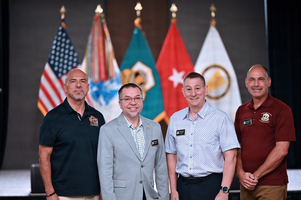Four men are posing together for a formal group photograph in front of a display of military flags, including the United States flag and several branch/unit flags. The setting appears to be an official military or government venue, with the individuals wearing a mix of business casual and polo-style attire with name badges. The image suggests an official event, ceremony, or conference.