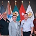 Four men are posing together for a formal group photograph in front of a display of military flags, including the United States flag and several branch/unit flags. The setting appears to be an official military or government venue, with the individuals wearing a mix of business casual and polo-style attire with name badges. The image suggests an official event, ceremony, or conference.