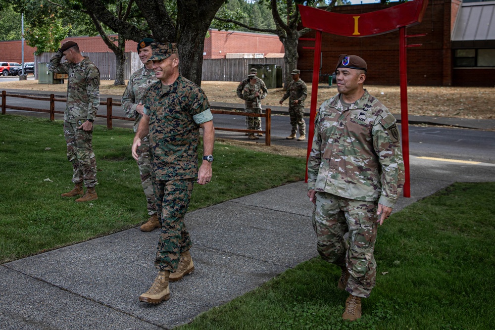 A group of military personnel from multiple branches are walking along a sidewalk on what appears to be a U.S. military installation. The foreground features a U.S. Marine in MARPAT camouflage alongside a U.S. Army soldier, with additional Army personnel visible in the background. A distinctive red torii-style gate with the Roman numeral 'I' is visible, suggesting this may be a I Corps installation such as Joint Base Lewis-McChord.