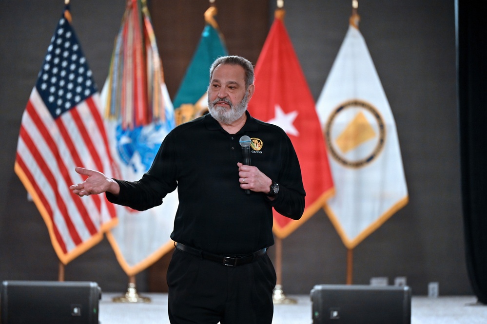 A middle-aged man with a gray beard is speaking at a formal event, holding a microphone and gesturing with his left hand. He is wearing a black polo shirt with an embroidered patch reading 'G6/CIO' on the chest. Several flags are displayed behind him, including the United States flag and what appear to be U.S. Army-related flags.