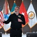 A middle-aged man with a gray beard is speaking at a formal event, holding a microphone and gesturing with his left hand. He is wearing a black polo shirt with an embroidered patch reading 'G6/CIO' on the chest. Several flags are displayed behind him, including the United States flag and what appear to be U.S. Army-related flags.