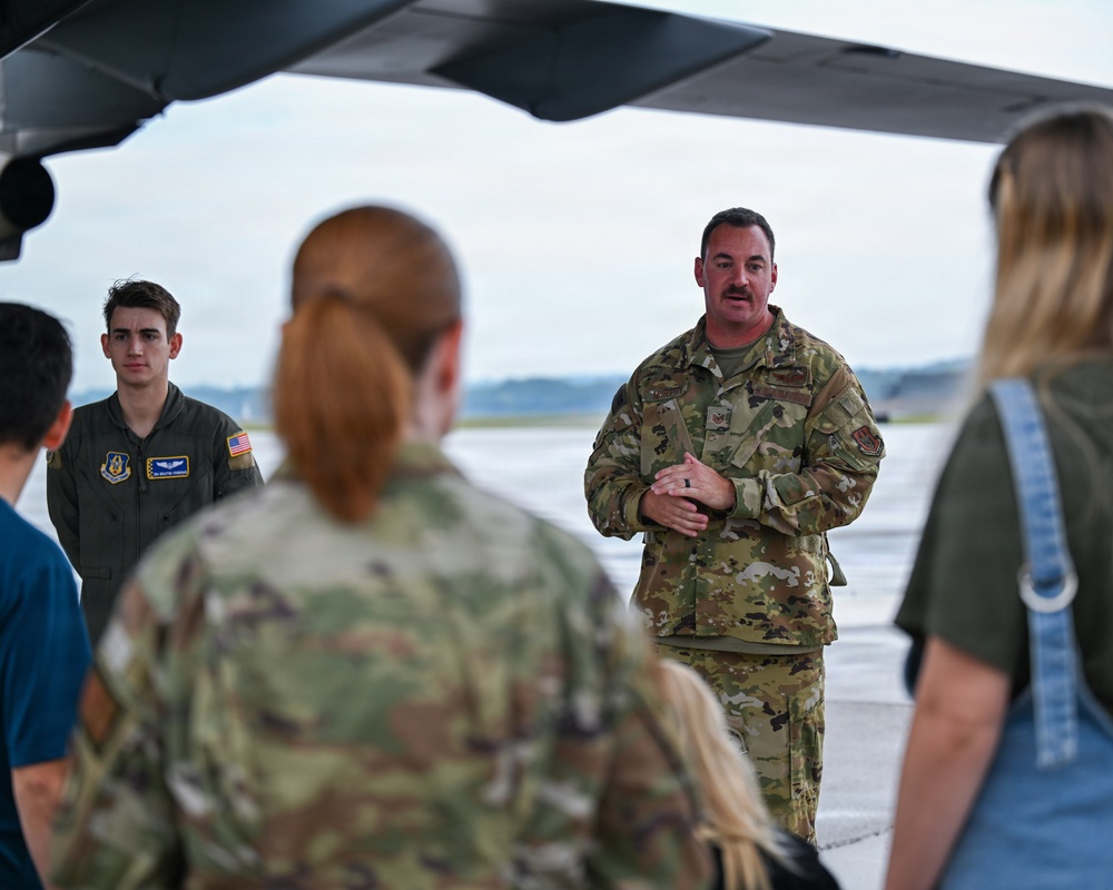 A military briefing or orientation is taking place on an airfield tarmac beneath the wing of a large aircraft. A senior military member in OCP (Operational Camouflage Pattern) uniform appears to be addressing a small group that includes both uniformed personnel and civilians. The setting suggests a tour, familiarization event, or unit visit at a military air base.