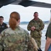 A military briefing or orientation is taking place on an airfield tarmac beneath the wing of a large aircraft. A senior military member in OCP (Operational Camouflage Pattern) uniform appears to be addressing a small group that includes both uniformed personnel and civilians. The setting suggests a tour, familiarization event, or unit visit at a military air base.