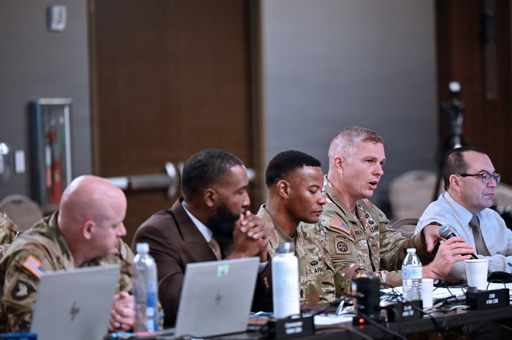 A panel discussion or briefing is taking place in what appears to be a conference room or event hall. Several U.S. Army personnel in camouflage uniforms are seated at a long table alongside civilians, with one soldier speaking into a microphone. The setting suggests a formal military meeting, symposium, or community engagement event.