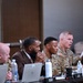 A panel discussion or briefing is taking place in what appears to be a conference room or event hall. Several U.S. Army personnel in camouflage uniforms are seated at a long table alongside civilians, with one soldier speaking into a microphone. The setting suggests a formal military meeting, symposium, or community engagement event.