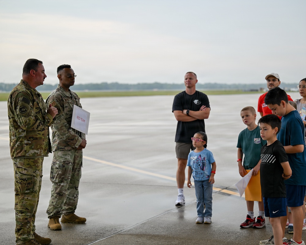 Two military personnel in camouflage uniforms are addressing a small group of civilians, including several children, on what appears to be an airfield tarmac. One of the soldiers is holding a sign labeled 'RED TEAM,' suggesting this is an organized activity or exercise involving families. The setting appears to be a military base open house, family day, or similar community engagement event.