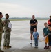 Two military personnel in camouflage uniforms are addressing a small group of civilians, including several children, on what appears to be an airfield tarmac. One of the soldiers is holding a sign labeled 'RED TEAM,' suggesting this is an organized activity or exercise involving families. The setting appears to be a military base open house, family day, or similar community engagement event.