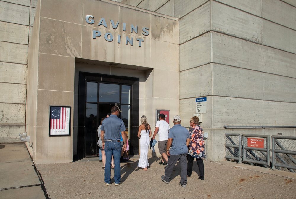 A group of visitors is entering the Gavins Point facility, which appears to be a dam or hydroelectric power station managed by the U.S. Army Corps of Engineers. The concrete building displays the name 'GAVINS POINT' in large letters above the entrance, along with a poster featuring a Betsy Ross-style flag reading '200 Years.' Several people of varying ages are walking toward the glass entrance doors.