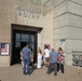 A group of visitors is entering the Gavins Point facility, which appears to be a dam or hydroelectric power station managed by the U.S. Army Corps of Engineers. The concrete building displays the name 'GAVINS POINT' in large letters above the entrance, along with a poster featuring a Betsy Ross-style flag reading '200 Years.' Several people of varying ages are walking toward the glass entrance doors.