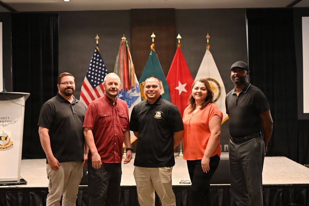 Five individuals pose together in front of a display of military and U.S. flags at what appears to be a formal event or ceremony. The setting includes a podium on the left with partial 'Marriott San Antonio' text visible, suggesting a hotel conference or banquet venue. The group appears to be associated with a military or government organization based on the insignia visible on one person's polo shirt.