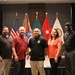 Five individuals pose together in front of a display of military and U.S. flags at what appears to be a formal event or ceremony. The setting includes a podium on the left with partial 'Marriott San Antonio' text visible, suggesting a hotel conference or banquet venue. The group appears to be associated with a military or government organization based on the insignia visible on one person's polo shirt.