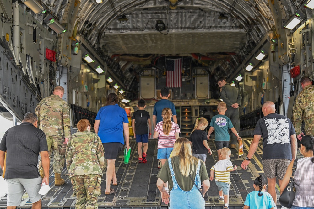 A group of civilians, including adults and children, are walking into the cargo hold of a large military transport aircraft, likely a C-17 Globemaster III. Several uniformed military personnel in camouflage (OCP/MultiCam) are present, appearing to guide or escort the visitors. An American flag is visible mounted at the far end of the cargo bay, suggesting this is a public tour or family day event at a military installation.