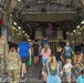 A group of civilians, including adults and children, are walking into the cargo hold of a large military transport aircraft, likely a C-17 Globemaster III. Several uniformed military personnel in camouflage (OCP/MultiCam) are present, appearing to guide or escort the visitors. An American flag is visible mounted at the far end of the cargo bay, suggesting this is a public tour or family day event at a military installation.