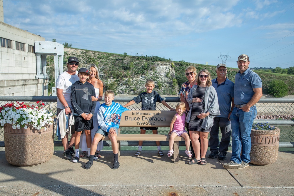 A group of approximately ten people, spanning multiple generations, are gathered around a memorial bench dedicated to Bruce Dvoracek (1999-2022). The photo appears to be a family memorial gathering at an outdoor public area, likely near a dam or waterway infrastructure. Two children are seated on the bench while adults stand around it, suggesting a commemorative family portrait.
