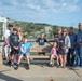 A group of approximately ten people, spanning multiple generations, are gathered around a memorial bench dedicated to Bruce Dvoracek (1999-2022). The photo appears to be a family memorial gathering at an outdoor public area, likely near a dam or waterway infrastructure. Two children are seated on the bench while adults stand around it, suggesting a commemorative family portrait.