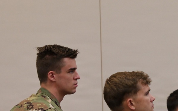 The image shows multiple military personnel in U.S. Army Operational Camouflage Pattern (OCP) uniforms seated in what appears to be a briefing or classroom setting. They are attentively looking forward, suggesting they are receiving instruction or attending a presentation. American flag patches are visible on their left shoulders, consistent with U.S. Army uniform regulations.