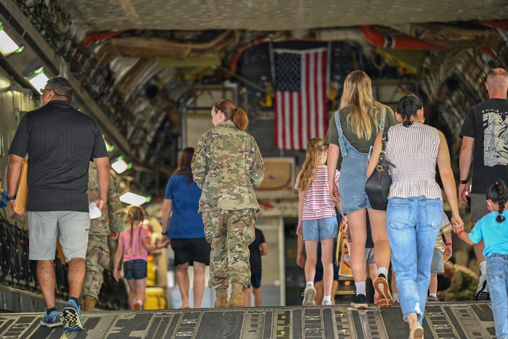 A group of adults and children are walking into the cargo hold of a large military transport aircraft, likely a C-17 Globemaster III. An American flag is displayed inside the aircraft's interior, suggesting this is a public open house, family day, or airshow event at a military installation. At least one uniformed soldier in Army Combat Uniform (ACU) is visible escorting or accompanying the civilian visitors.