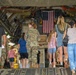 A group of adults and children are walking into the cargo hold of a large military transport aircraft, likely a C-17 Globemaster III. An American flag is displayed inside the aircraft's interior, suggesting this is a public open house, family day, or airshow event at a military installation. At least one uniformed soldier in Army Combat Uniform (ACU) is visible escorting or accompanying the civilian visitors.