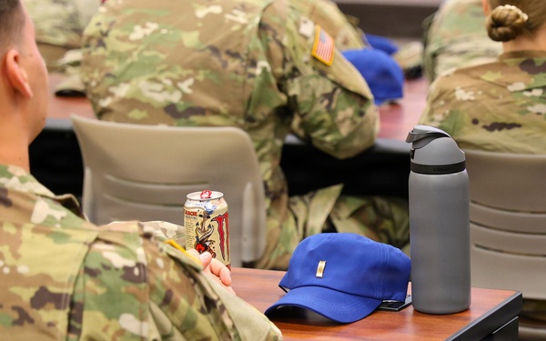 Several military personnel in U.S. Army OCP (Operational Camouflage Pattern) uniforms are seated in what appears to be a classroom or briefing room setting. The foreground soldier holds an energy drink can and has a blue baseball cap and gray water bottle on the desk. American flag patches are visible on the uniforms, indicating active U.S. Army service members.