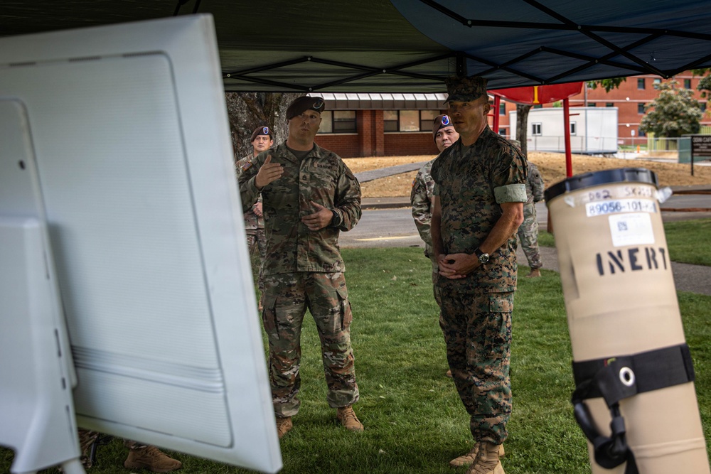 Military personnel in camouflage uniforms are gathered outdoors under a blue canopy tent, appearing to conduct a briefing or demonstration. An Army soldier appears to be presenting or explaining something to a Marine Corps officer and other personnel. A cylindrical object labeled 'INERT' is visible in the foreground, indicating a training exercise involving inert ordnance or equipment.