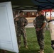 Military personnel in camouflage uniforms are gathered outdoors under a blue canopy tent, appearing to conduct a briefing or demonstration. An Army soldier appears to be presenting or explaining something to a Marine Corps officer and other personnel. A cylindrical object labeled 'INERT' is visible in the foreground, indicating a training exercise involving inert ordnance or equipment.