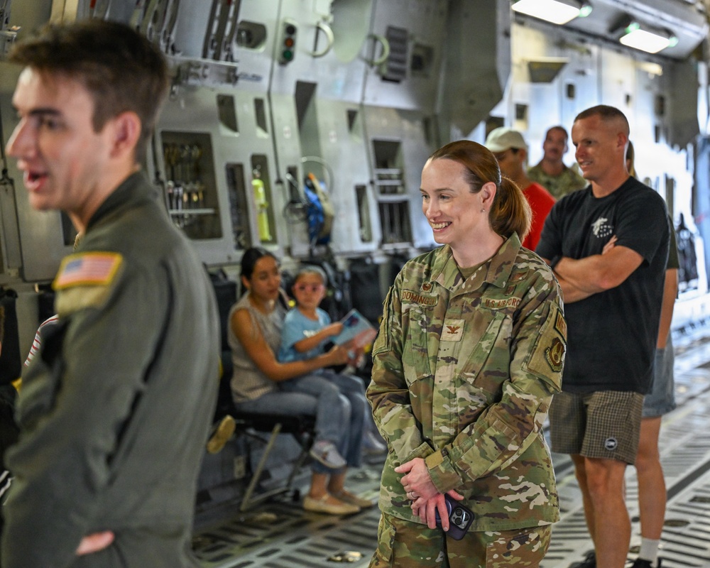 The image shows a group of people inside what appears to be a large military transport aircraft, likely a C-17 Globemaster III, based on the interior cargo bay features. A U.S. Air Force officer in OCP (Operational Camouflage Pattern) uniform, with a colonel's eagle insignia and a name tape reading 'DOMINGUEZ,' is smiling and engaged in conversation. The setting appears to be an open house, tour, or family day event given the presence of civilians including children.