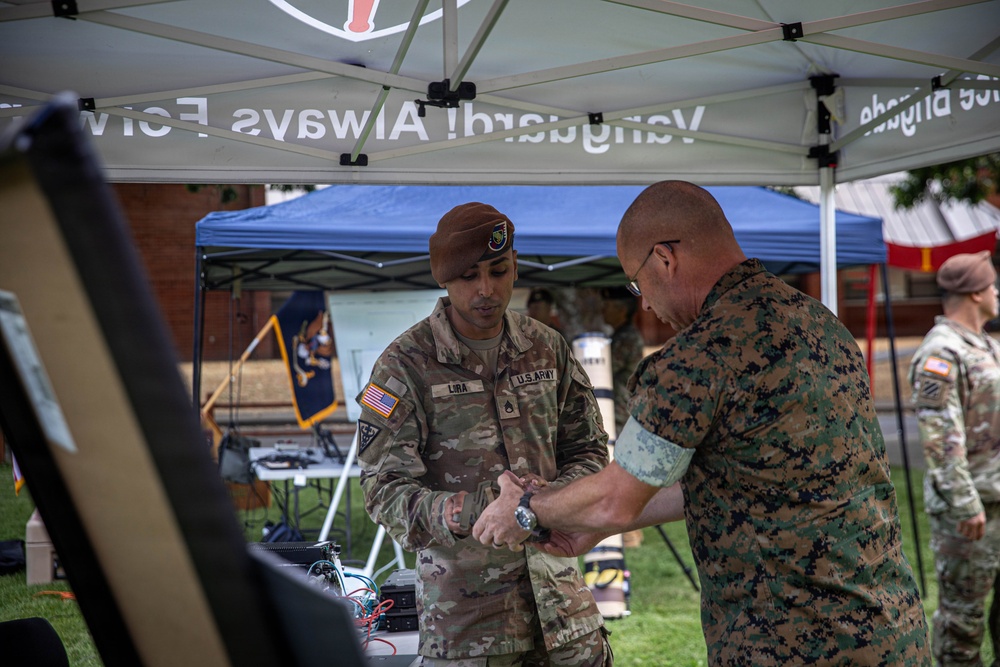 Two military personnel are interacting under a white canopy tent at what appears to be an outdoor military event or demonstration. A U.S. Army soldier wearing a brown beret and name tape reading 'LIRA' is working with a Marine in MARPAT camouflage, appearing to apply or adjust something on the Marine's wrist or arm. Additional military personnel are visible in the background under a blue canopy.