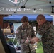 Two military personnel are interacting under a white canopy tent at what appears to be an outdoor military event or demonstration. A U.S. Army soldier wearing a brown beret and name tape reading 'LIRA' is working with a Marine in MARPAT camouflage, appearing to apply or adjust something on the Marine's wrist or arm. Additional military personnel are visible in the background under a blue canopy.