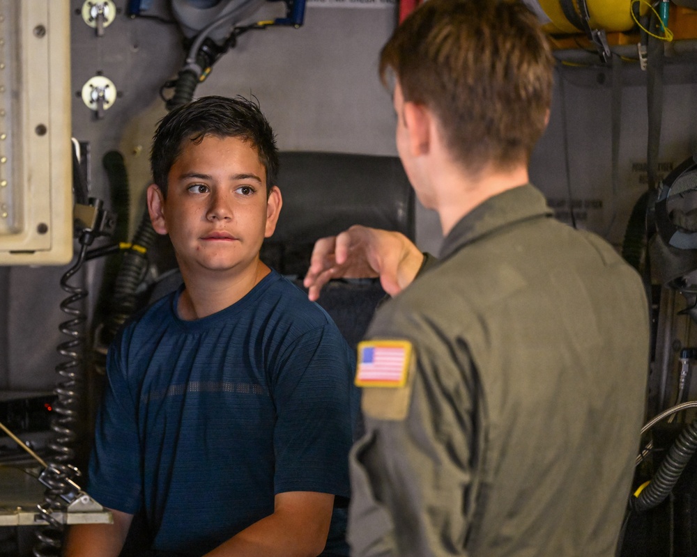 A young civilian boy in a blue t-shirt is speaking with a military personnel member wearing an olive green flight suit with a U.S. flag patch on the left shoulder. They appear to be inside a military aircraft or vehicle, surrounded by equipment, cables, and mechanical components. The interaction suggests a tour or educational visit to a military asset.