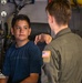 A young civilian boy in a blue t-shirt is speaking with a military personnel member wearing an olive green flight suit with a U.S. flag patch on the left shoulder. They appear to be inside a military aircraft or vehicle, surrounded by equipment, cables, and mechanical components. The interaction suggests a tour or educational visit to a military asset.