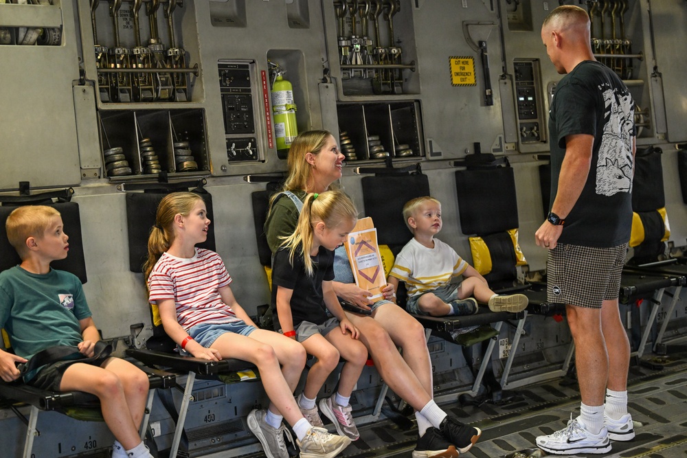 A group of children and an adult woman are seated on military aircraft jump seats inside what appears to be a large military transport aircraft, likely a C-17 Globemaster III. A man in civilian clothing stands facing them, appearing to give a tour or briefing. The setting suggests a public open house or family day event at a military installation.