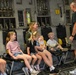 A group of children and an adult woman are seated on military aircraft jump seats inside what appears to be a large military transport aircraft, likely a C-17 Globemaster III. A man in civilian clothing stands facing them, appearing to give a tour or briefing. The setting suggests a public open house or family day event at a military installation.