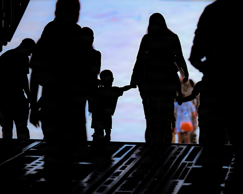 A silhouetted scene showing a group of people, including at least one small child holding hands with an adult, walking toward a bright opening or exit. The image is backlit, creating strong dark silhouettes against a blue and white luminous background. The flooring appears to be a structured metal ramp or platform, suggesting an industrial or transport setting.