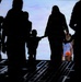 A silhouetted scene showing a group of people, including at least one small child holding hands with an adult, walking toward a bright opening or exit. The image is backlit, creating strong dark silhouettes against a blue and white luminous background. The flooring appears to be a structured metal ramp or platform, suggesting an industrial or transport setting.