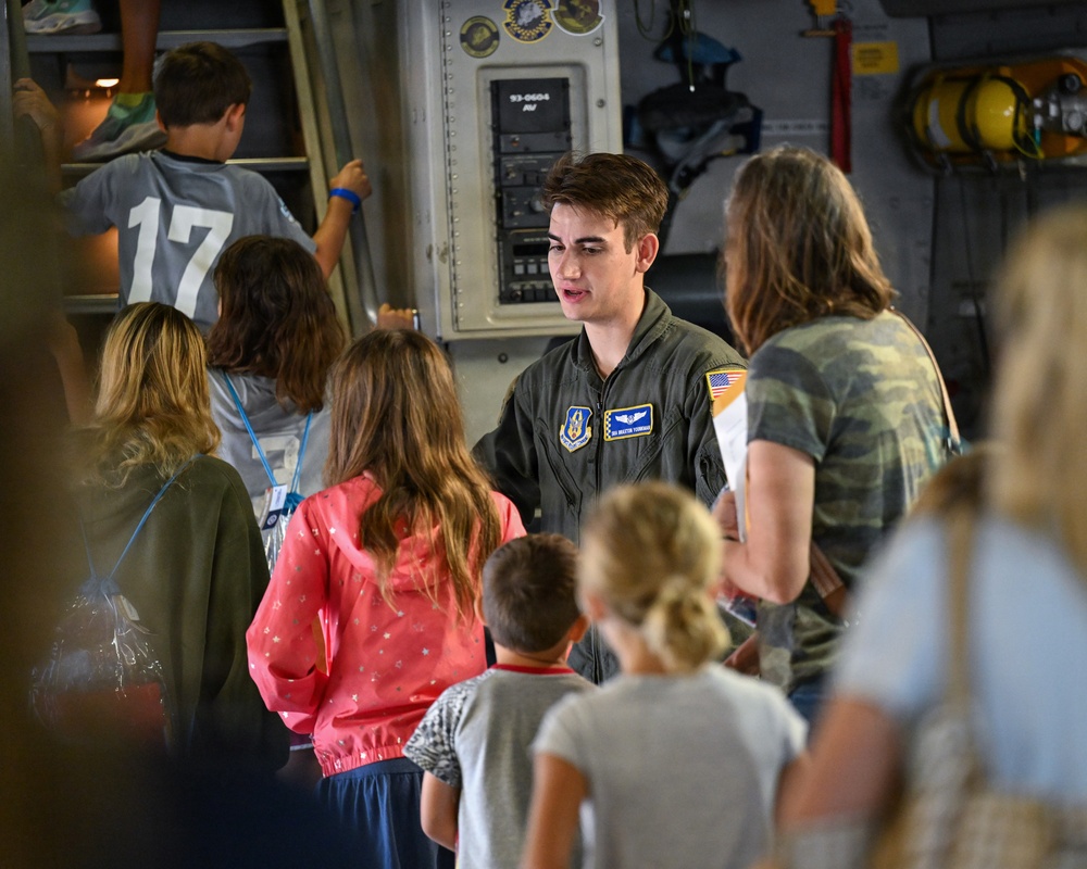 A young U.S. Air Force airman in a flight suit is conducting a tour or briefing for a group of children and adults inside a military aircraft. The airman appears to be explaining equipment or systems to the visitors, who are gathered around attentively. The setting appears to be an open house or public outreach event aboard a military transport aircraft.