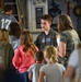 A young U.S. Air Force airman in a flight suit is conducting a tour or briefing for a group of children and adults inside a military aircraft. The airman appears to be explaining equipment or systems to the visitors, who are gathered around attentively. The setting appears to be an open house or public outreach event aboard a military transport aircraft.