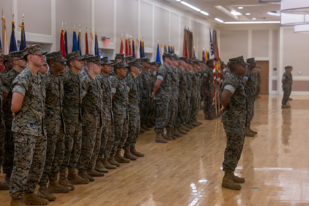 A formal military ceremony is taking place inside a large indoor hall with a polished hardwood floor. A large formation of U.S. Marines in digital camouflage (MARPAT) uniforms stands at attention or parade rest, with multiple unit and national flags displayed along the back wall. A senior non-commissioned officer or officer appears to be inspecting or addressing the formation from the front.