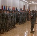 A formal military ceremony is taking place inside a large indoor hall with a polished hardwood floor. A large formation of U.S. Marines in digital camouflage (MARPAT) uniforms stands at attention or parade rest, with multiple unit and national flags displayed along the back wall. A senior non-commissioned officer or officer appears to be inspecting or addressing the formation from the front.
