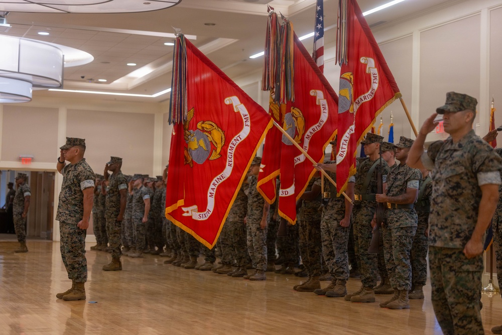 A formal U.S. Marine Corps ceremony is taking place indoors in what appears to be a ballroom or event hall. Multiple Marines in MARPAT camouflage uniforms are standing at attention and saluting while color guard members hold large red regimental and battalion guidons. The flags visible include those of the 6th Marine Regiment FMF and 3rd Battalion, along with a U.S. national flag.