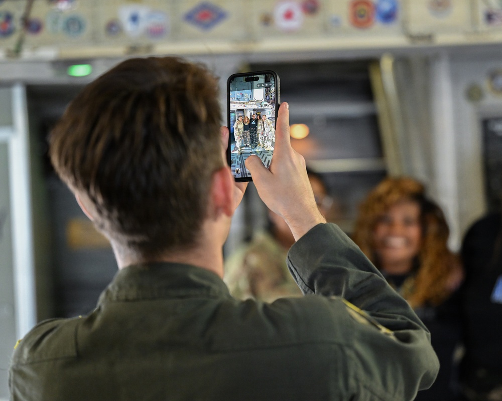 A person wearing an olive/military green flight suit is photographing a group of people using a smartphone inside what appears to be a military aircraft. The phone screen displays a group photo of several individuals in military uniforms. Colorful unit patches or insignia are visible along the upper interior wall of the aircraft.