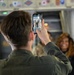 A person wearing an olive/military green flight suit is photographing a group of people using a smartphone inside what appears to be a military aircraft. The phone screen displays a group photo of several individuals in military uniforms. Colorful unit patches or insignia are visible along the upper interior wall of the aircraft.