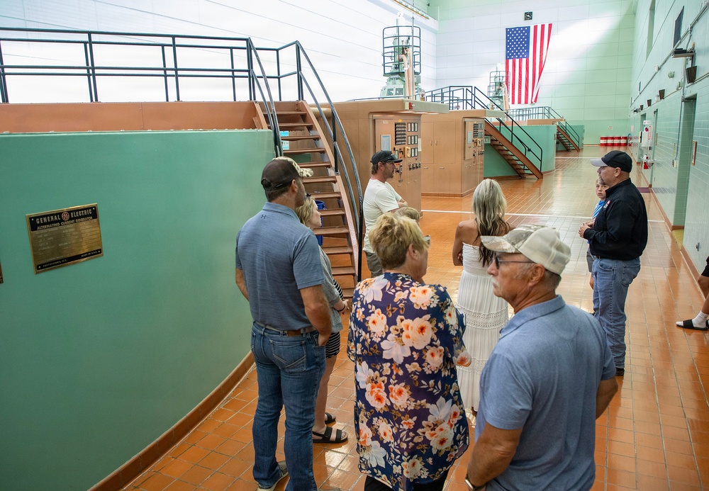 A group of visitors is touring what appears to be a hydroelectric power plant or industrial facility. They are gathered around large green-painted generator equipment bearing a 'General Electric Alternating Current Generator' nameplate. An American flag is prominently displayed in the background of the large industrial hall.