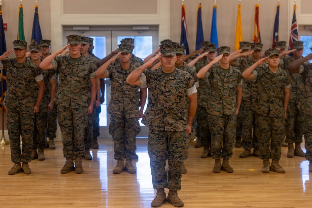 A group of U.S. Marines in MARPAT woodland camouflage uniforms are standing at attention and rendering a salute during an indoor military ceremony. The formation is led by a senior Marine in the foreground, with rows of personnel arranged behind in a formal indoor venue. Multiple state and international flags are displayed along the back wall.