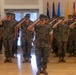 A group of U.S. Marines in MARPAT woodland camouflage uniforms are standing at attention and rendering a salute during an indoor military ceremony. The formation is led by a senior Marine in the foreground, with rows of personnel arranged behind in a formal indoor venue. Multiple state and international flags are displayed along the back wall.