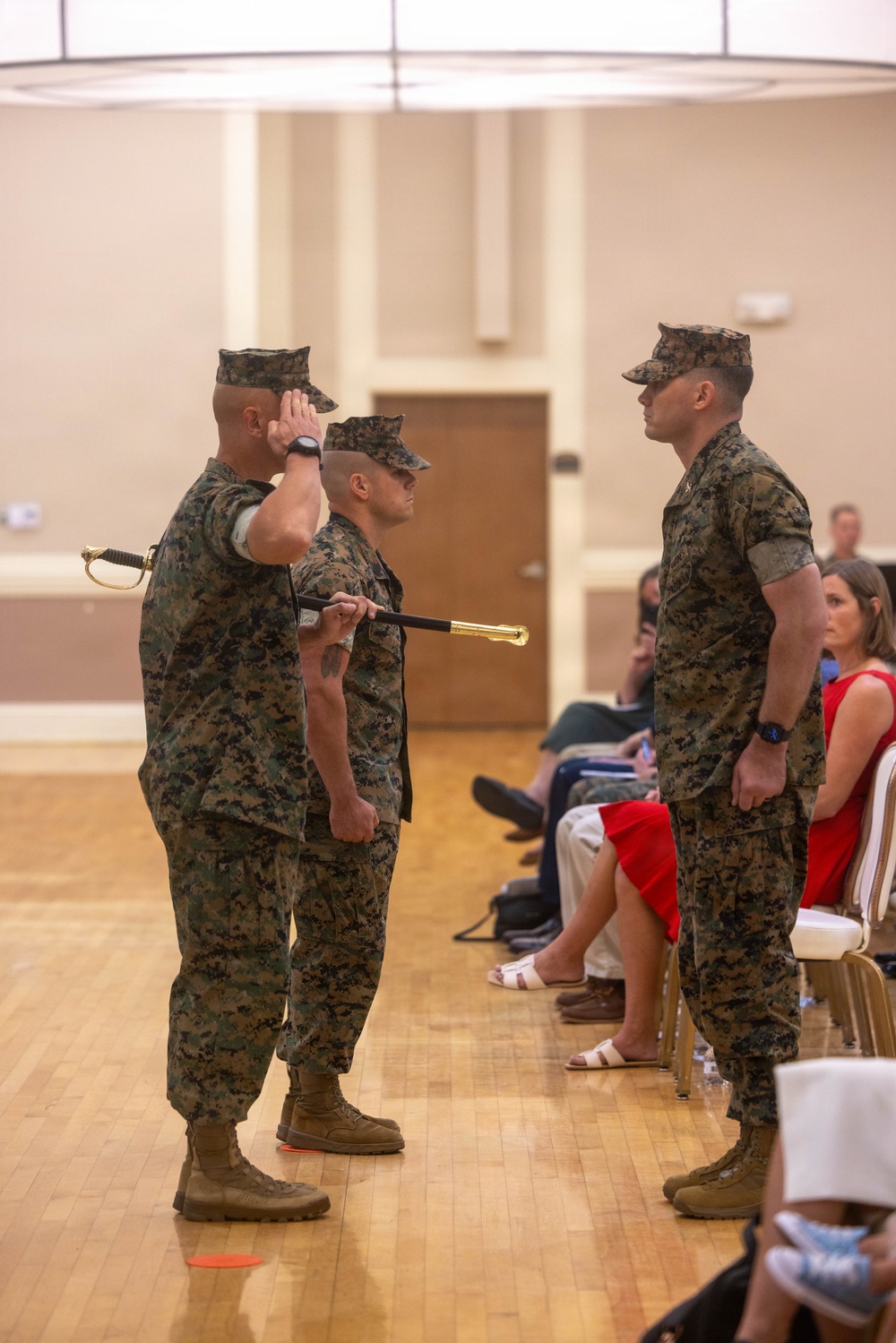A formal U.S. Marine Corps military ceremony is taking place indoors in what appears to be a community or event hall. Three Marines in MARPAT digital camouflage uniforms are participating in a ceremony, with one rendering a salute and another holding a ceremonial sword and a baton or staff. Seated civilians, likely family members, observe the proceedings in the background.