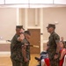 A formal U.S. Marine Corps military ceremony is taking place indoors in what appears to be a community or event hall. Three Marines in MARPAT digital camouflage uniforms are participating in a ceremony, with one rendering a salute and another holding a ceremonial sword and a baton or staff. Seated civilians, likely family members, observe the proceedings in the background.