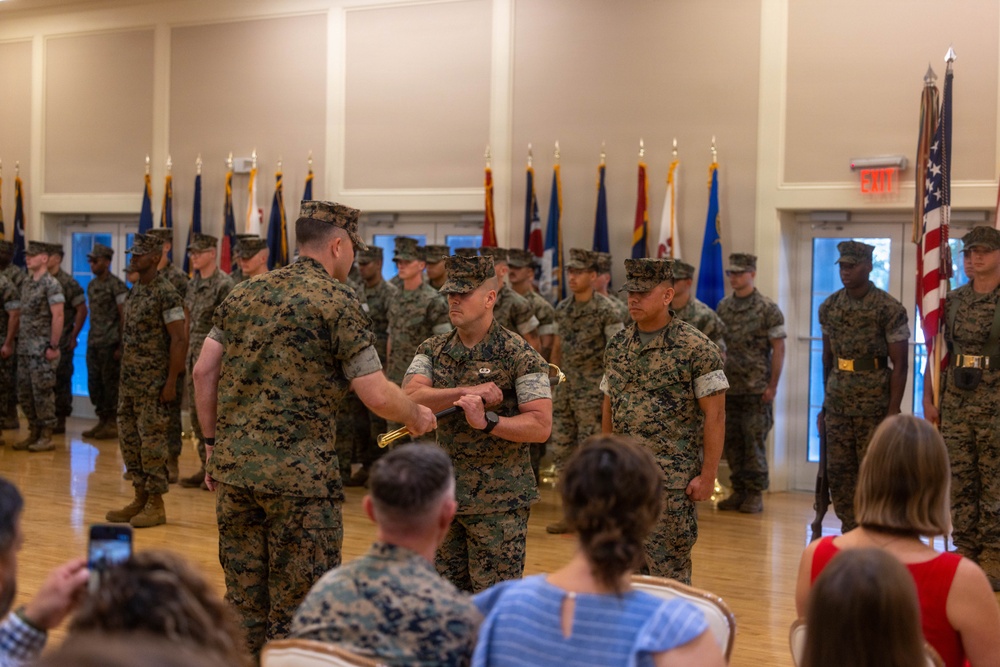 A formal U.S. Marine Corps military ceremony is taking place in an indoor hall, likely a change of command or awards ceremony. Several Marines in MARPAT camouflage uniforms are participating in a central exchange, possibly involving a ceremonial item such as a guidon or sword. Civilians and additional service members are seated and standing as observers.