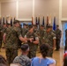 A formal U.S. Marine Corps military ceremony is taking place in an indoor hall, likely a change of command or awards ceremony. Several Marines in MARPAT camouflage uniforms are participating in a central exchange, possibly involving a ceremonial item such as a guidon or sword. Civilians and additional service members are seated and standing as observers.