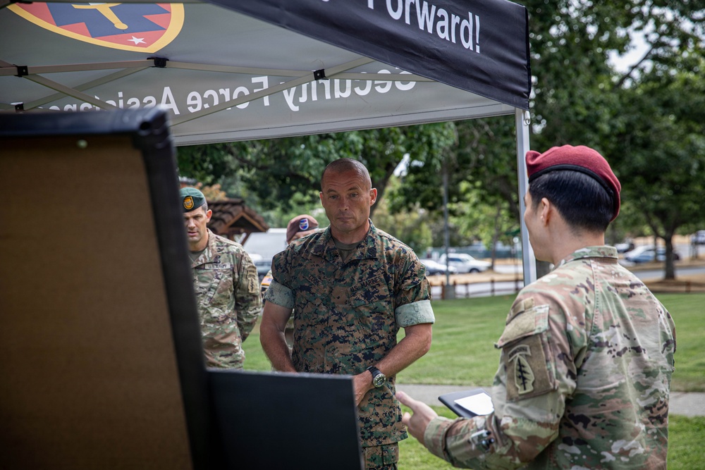 Military personnel from multiple branches are gathered under a black canopy tent at an outdoor event. A U.S. Marine in MARPAT camouflage is being briefed or presented to by a soldier wearing a maroon beret, while another soldier in a green beret stands in the background. The tent banner reads 'Security Force Assistance' and 'Forward!'